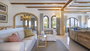 A spacious living area with a white sofa, a small side table, and a wooden cabinet. There are large windows with curtains, a wooden ceiling, and a stone archway. - Villa Pakatoa (Javea / Xàbia)