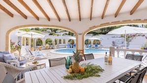 A poolside area with lounge chairs, a dining table with fruits and drinks, and a view of a pool and garden. - Villa Pakatoa (Javea / Xàbia)