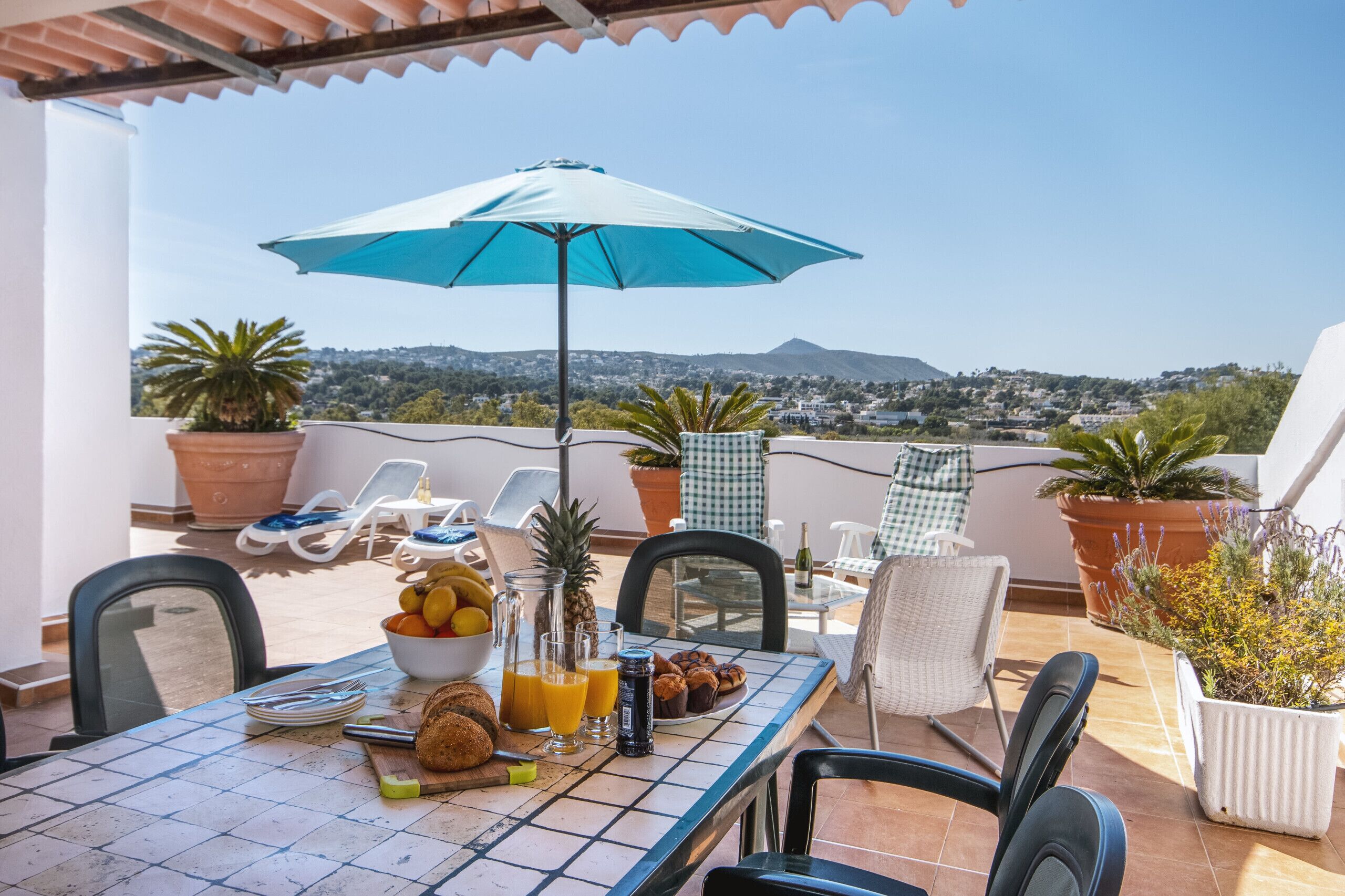A terrace with a table set for breakfast, including fruits, pastries, and drinks, with a view of a cityscape and mountains in the background.