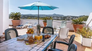 A terrace with a table set for breakfast, including fruits, pastries, and drinks, with a view of a cityscape and mountains in the background. - Atico Salonica (Javea / Xàbia)