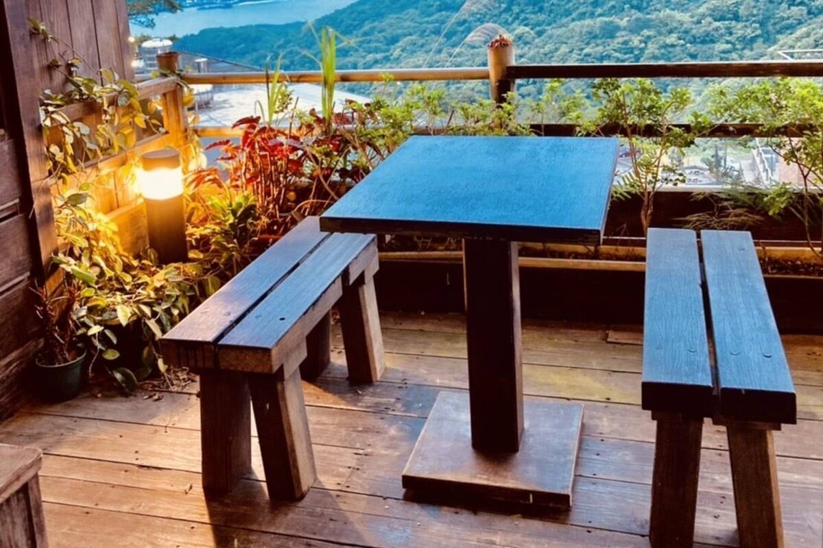 An outdoor patio with wooden benches and a table, surrounded by plants and a view of a mountainous landscape.