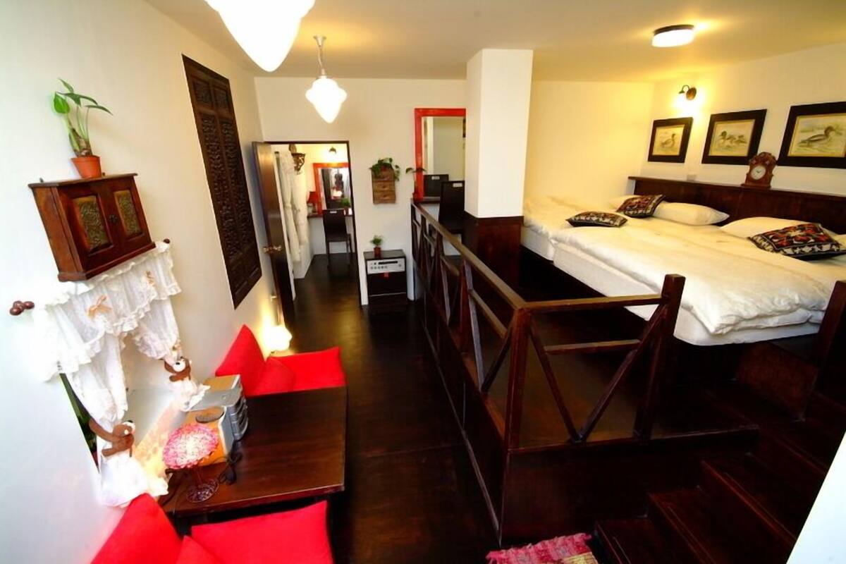 A compact hotel room with a bed, a small table, and a chair. There is a staircase leading to an upper level.