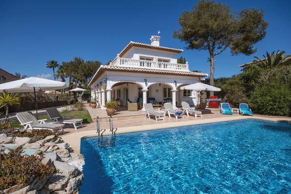Un área junto a la piscina con sillones de descanso, sombrillas y una piscina azul cristalina.