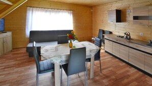 Una cabaña acogedora de madera con comedor, que incluye una mesa redonda, sillas y un jarrón con flores. Se ve una kitchenette con armarios y una estufa.