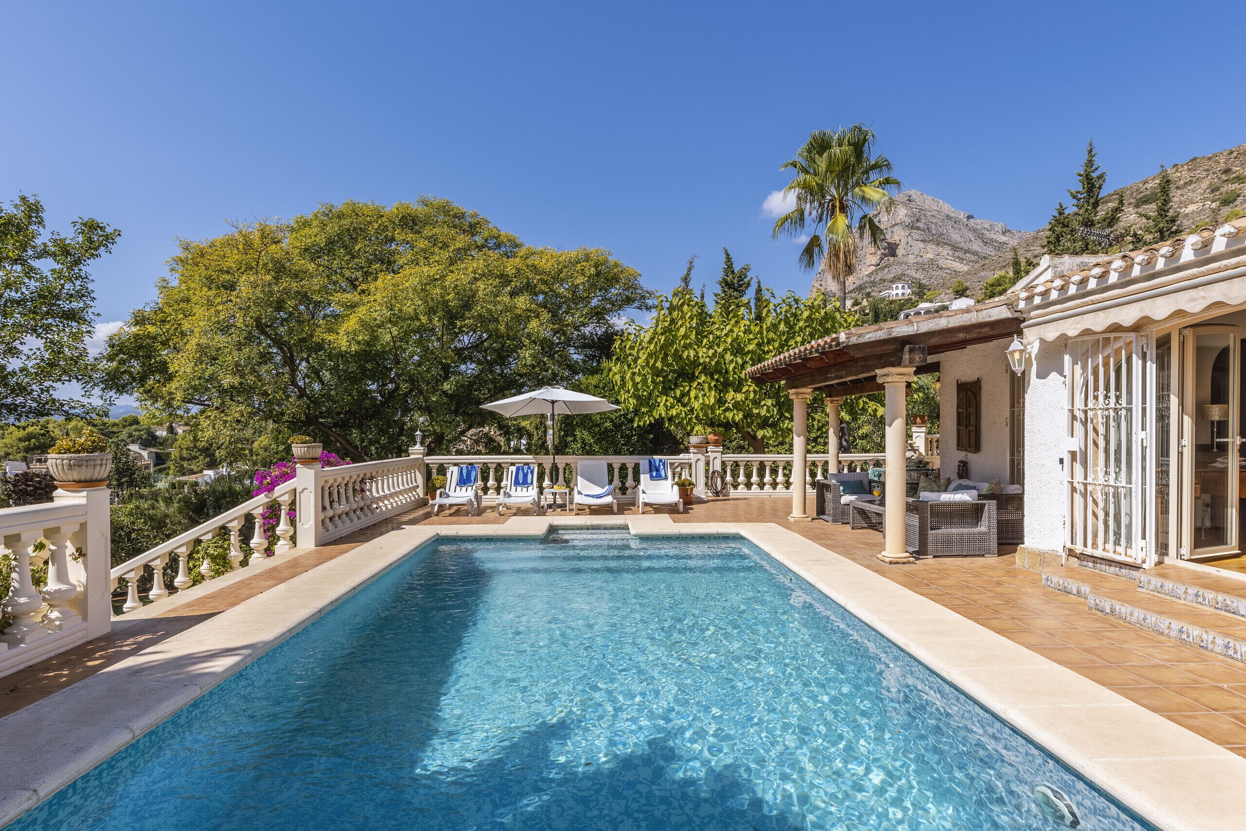 A rectangular swimming pool with a white balustrade, surrounded by lounge chairs and umbrellas, with a mountain and trees in the background.
