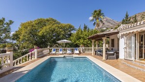 A rectangular swimming pool with a white balustrade, surrounded by lounge chairs and umbrellas, with a mountain and trees in the background. - Villa Levante (Javea / Xàbia)