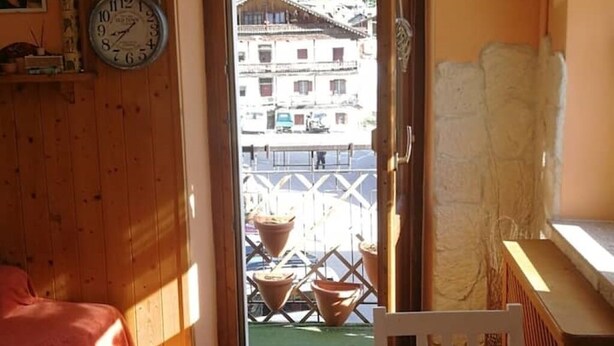 A room with a wooden floor, a bed with a pink blanket, a white chair, a wooden door, and a view of a building with balconies and a fence. - Relaxation & Skiing in the Heart of the Dolomites UNESCO (Calalzo di Cadore)