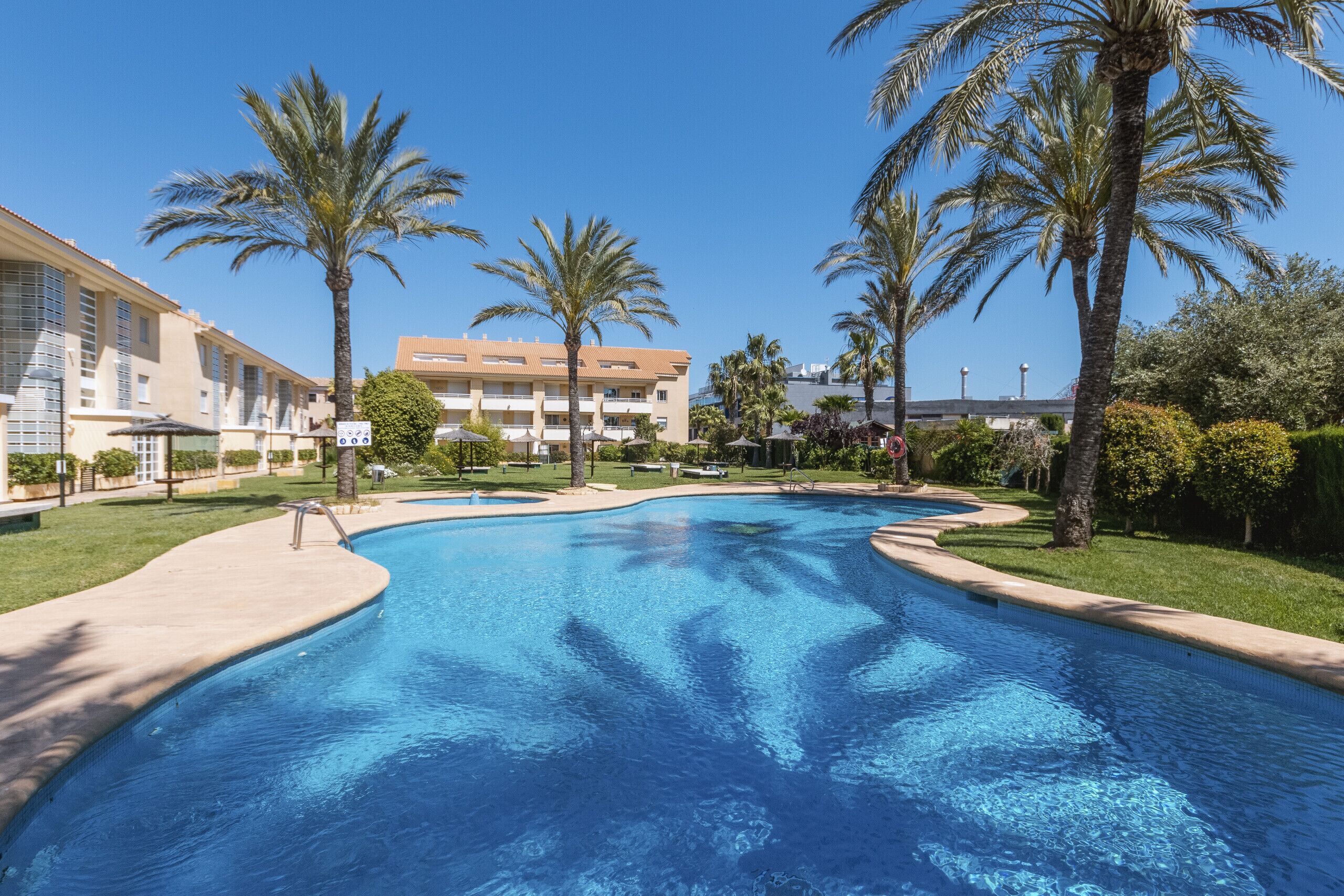 A swimming pool with palm trees, a building with balconies, and a clear blue sky.