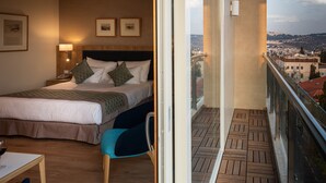 A hotel room with a bed, a chair, a table with a glass of wine, and a balcony with a city view. - Ambassador Boutique Hotel (Jerusalem)