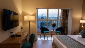 A hotel room with a bed, a desk, two chairs, a TV, a window with a city view, and a lamp. - Ambassador Boutique Hotel (Jerusalem)