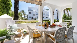 Une terrasse meublée de blanc, une table dressée avec de la nourriture et des boissons, et une vue sur des bâtiments blancs.