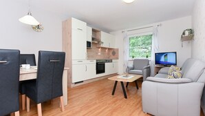 Moderni olohuone, jossa on harmaa sohva, ruokapöytä tuoleineen, televisio sekä keittiöalue, jossa on puukalusteet ja mikroaaltouuni.