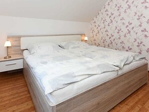 A bedroom with a wooden bed frame, white bedding, and a nightstand with a lamp.