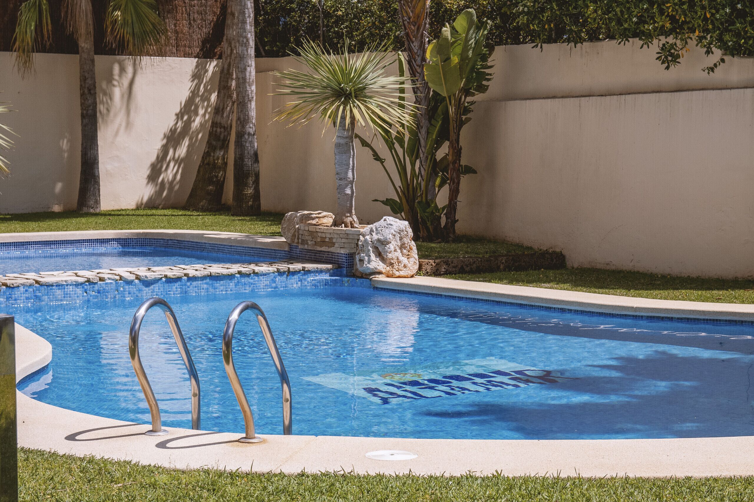 A swimming pool with clear blue water, a ladder, and a small rock feature.