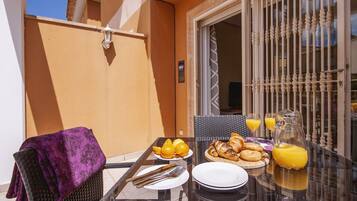 A breakfast table with croissants, orange juice, and a pitcher, set on a balcony with a wicker chair and a purple throw.