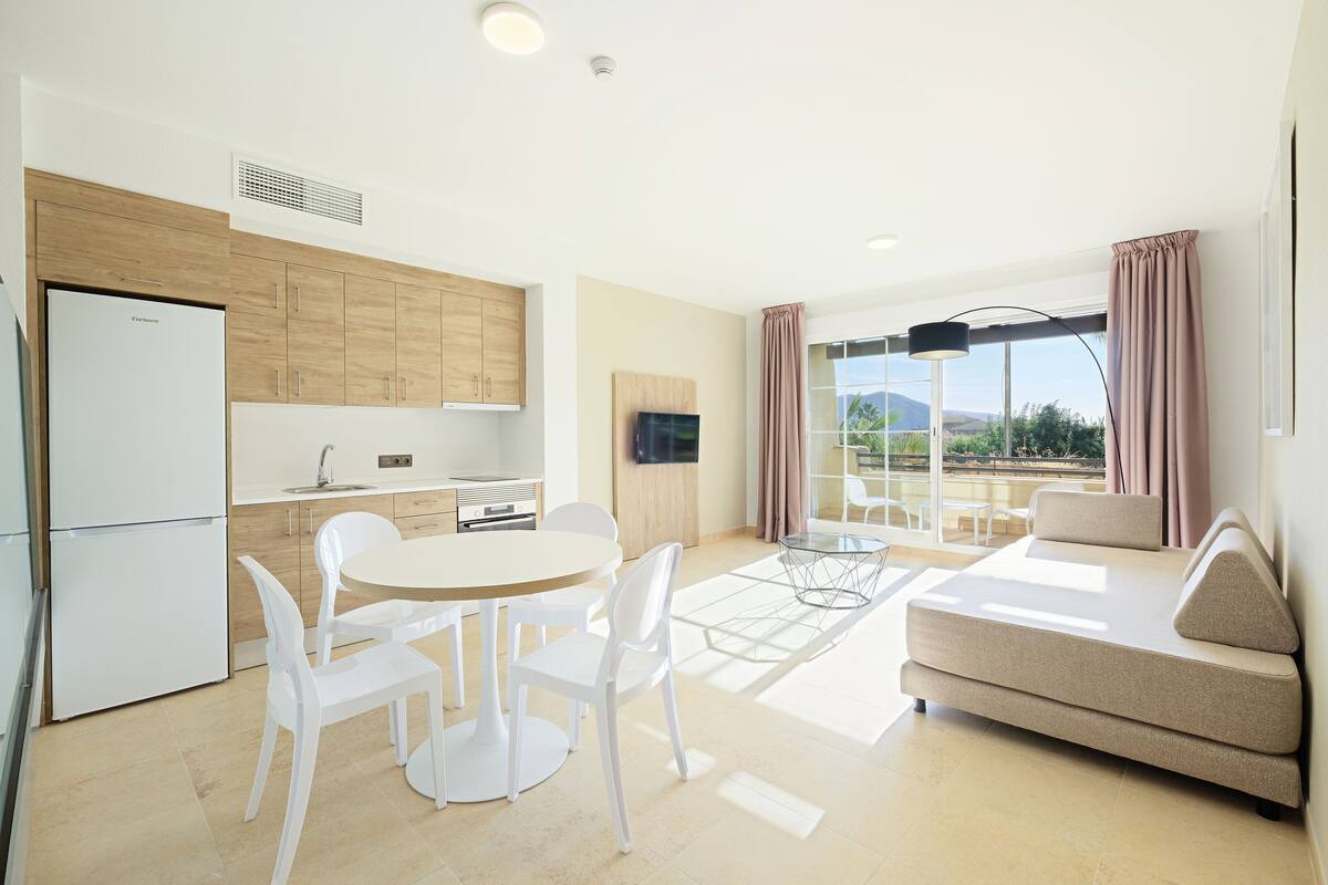 A modern living room with a sofa, a dining table with chairs, and a kitchen area with cabinets and a refrigerator.