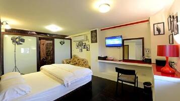 A hotel room with a bed, a chair, a TV, and a red lamp.