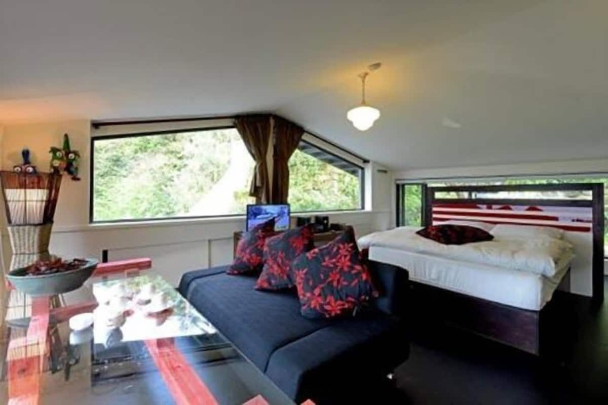 A modern room with a glass dining table, a sofa with red and black patterned pillows, a bed with a white headboard, and a large window with curtains.