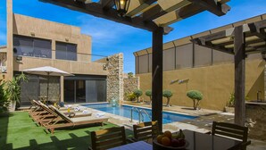 Zona de piscina con sillones de descanso, una mesa con fruta y vista a un edificio con pared de piedra y un árbol.