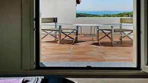 A view through a window of a terrace with a table and chairs, a view of the sea, and a building with a sign. - Apartment in Golfo Aranci with elevator (Baja Sardinia (SS))