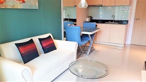 A compact living space with a white sofa, two blue chairs, a glass coffee table, and a kitchenette in the background. - Grande Caribbean Resort - Sleeps 2 (Pattaya)