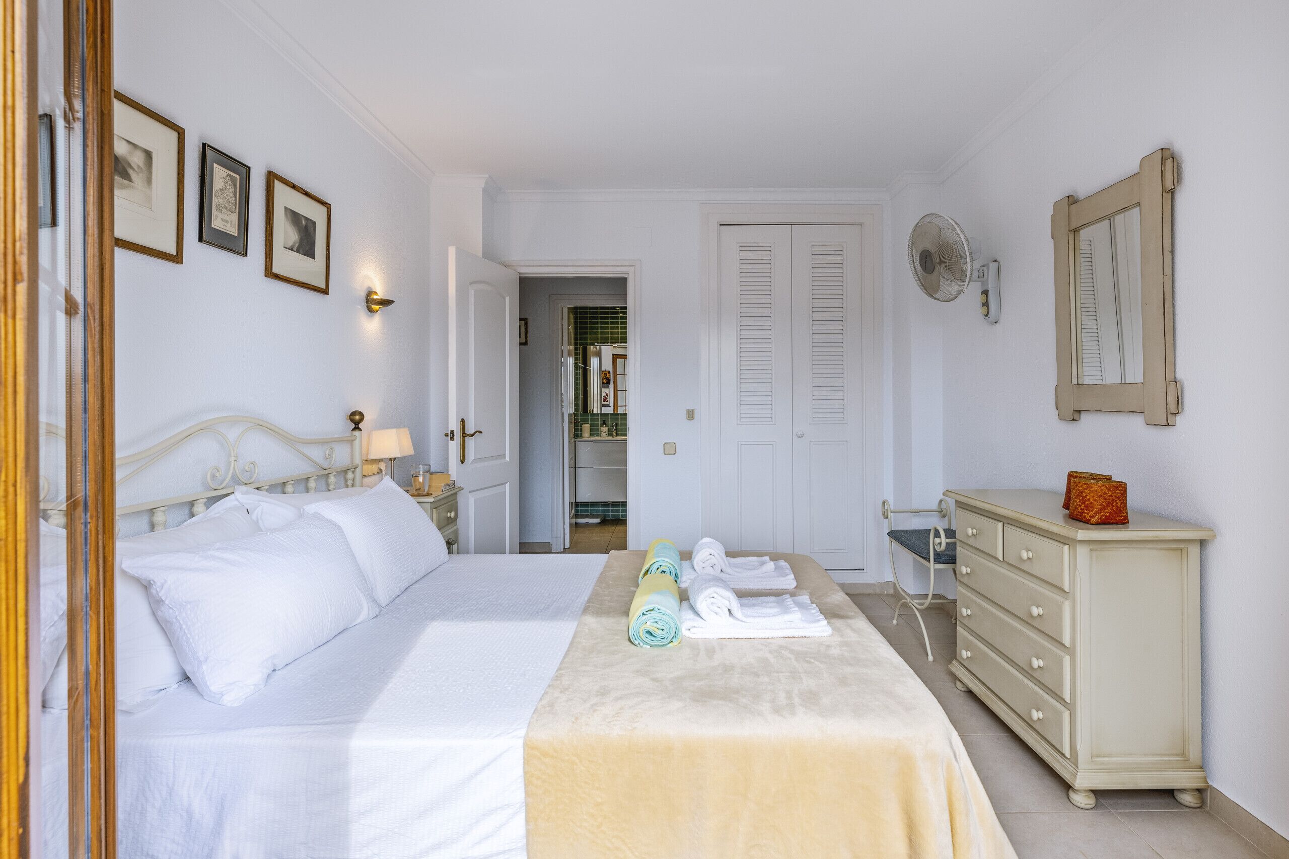 A bedroom with a bed, a dresser, and a mirror. There are towels on the dresser and a fan on the wall.