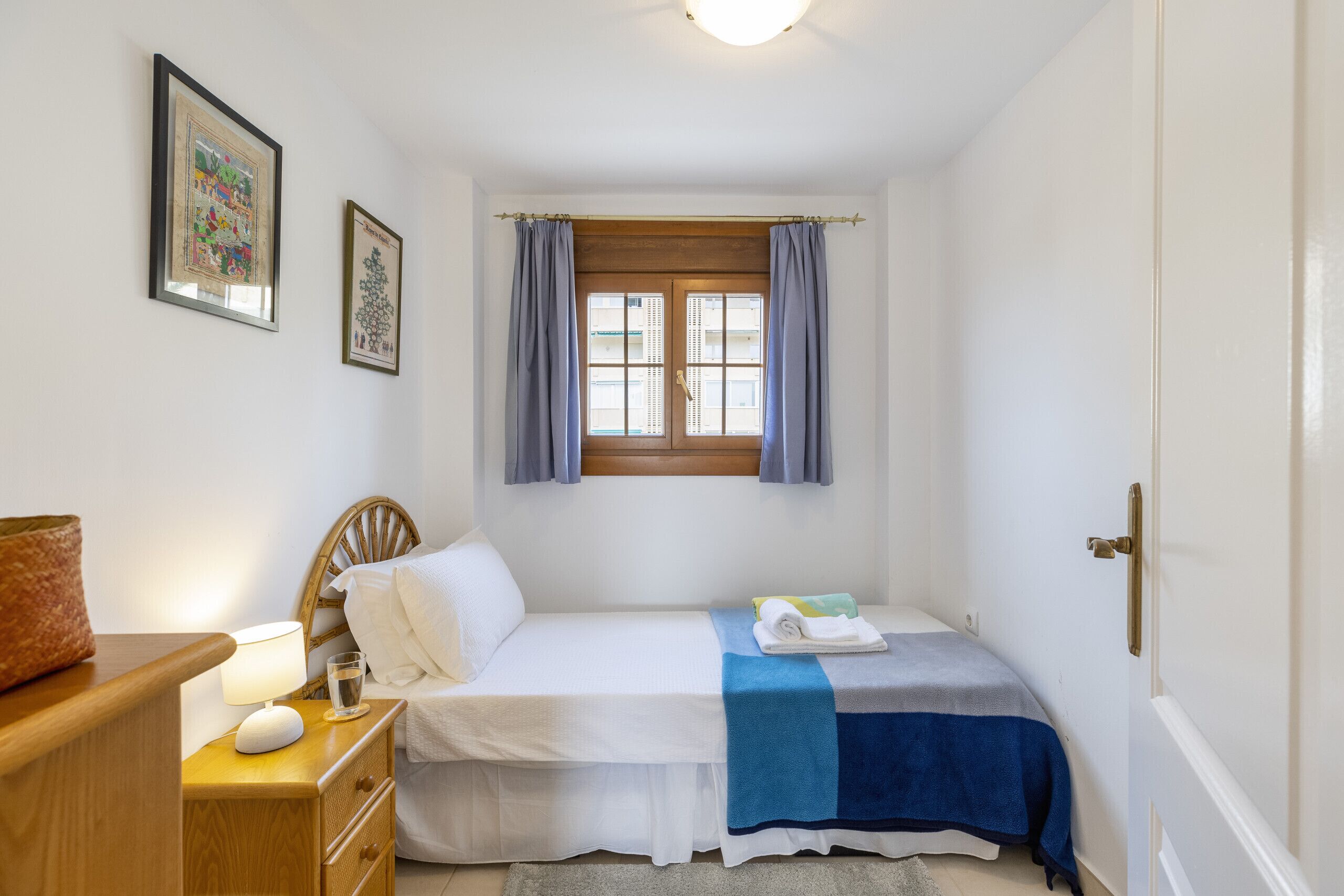 A small, neatly arranged bedroom with a single bed, a bedside table with a lamp, a window with curtains, and a door.