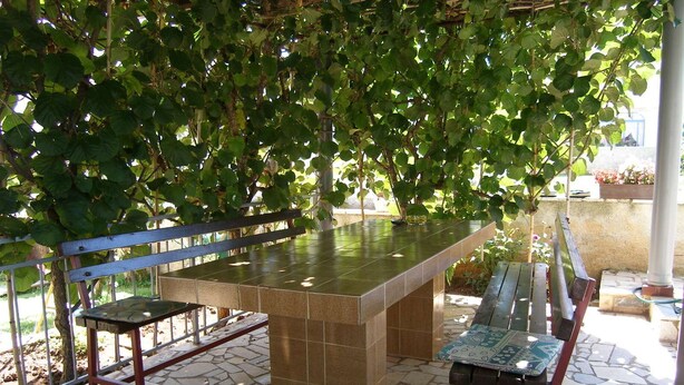 An outdoor seating area with a stone table and metal chairs, surrounded by greenery and stone flooring. - Apartments Violeta (Liznjan)