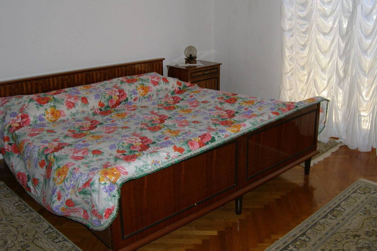 A bedroom with a wooden bed, floral bedspread, a small bedside table, and a white curtain.