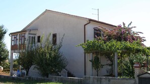 A single-story building with a flat roof, a balcony on the left side, and a small garden area with a tree and flowering plants. - Apartments Violeta (Liznjan)