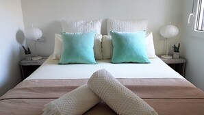 Una cama bien tendida con dos almohadas blancas y dos almohadas color turquesa, un plaid blanco y una manta blanca con textura. Hay dos mesitas de noche con lámparas y pequeñas macetas.