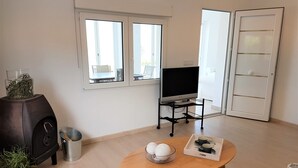 Una sala de estar con un televisor sobre un mueble bajo, una mesa de madera con adornos, una parrilla y una ventana con vistas.