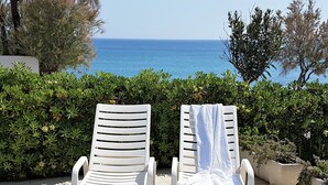 Dos sillones blancos con toallas, ubicados en un patio con vista al mar y un seto al fondo.