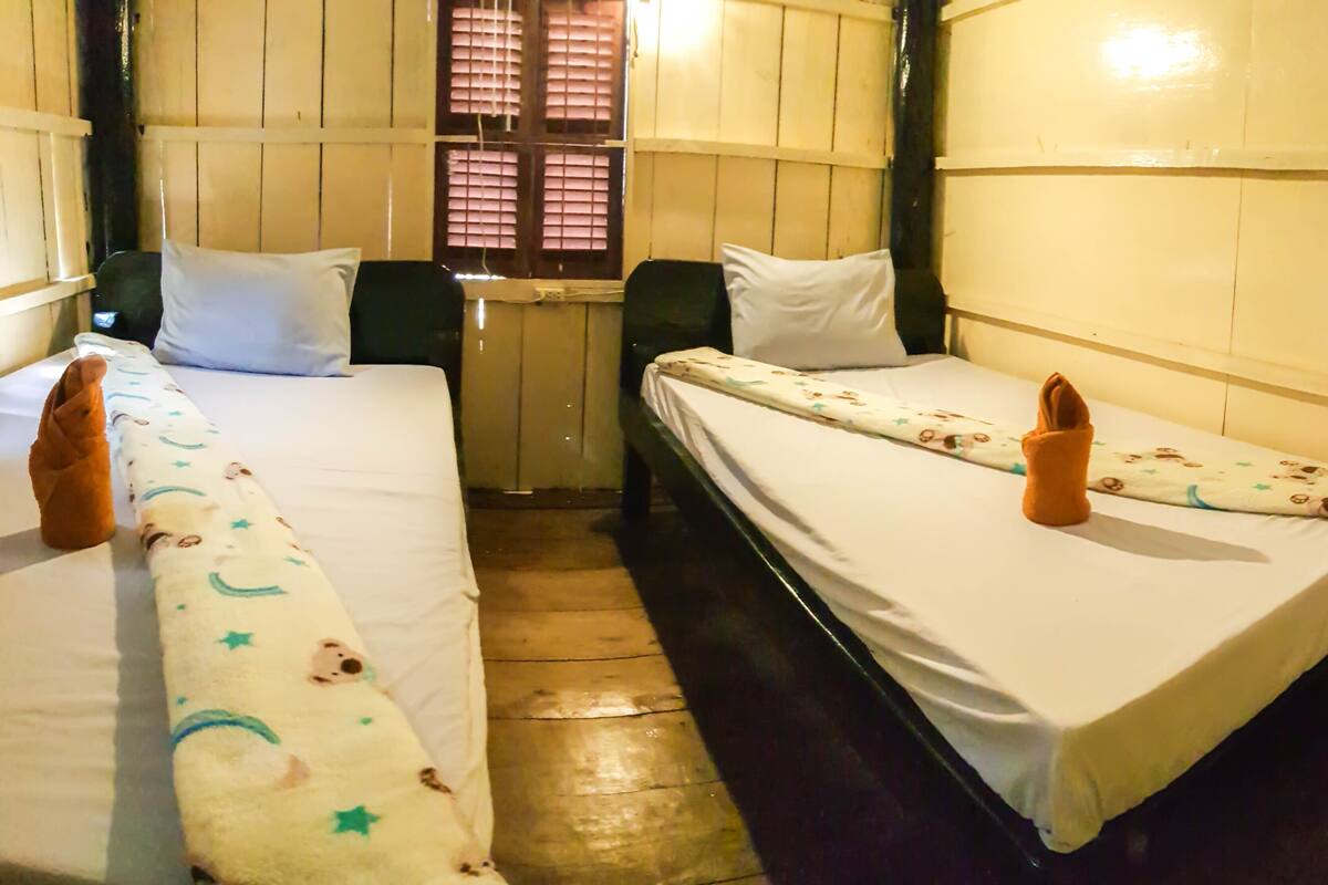 A room with two single beds, each with a pillow and a folded towel. The beds have white linens with a pattern. The room has wooden walls and a window with blinds.