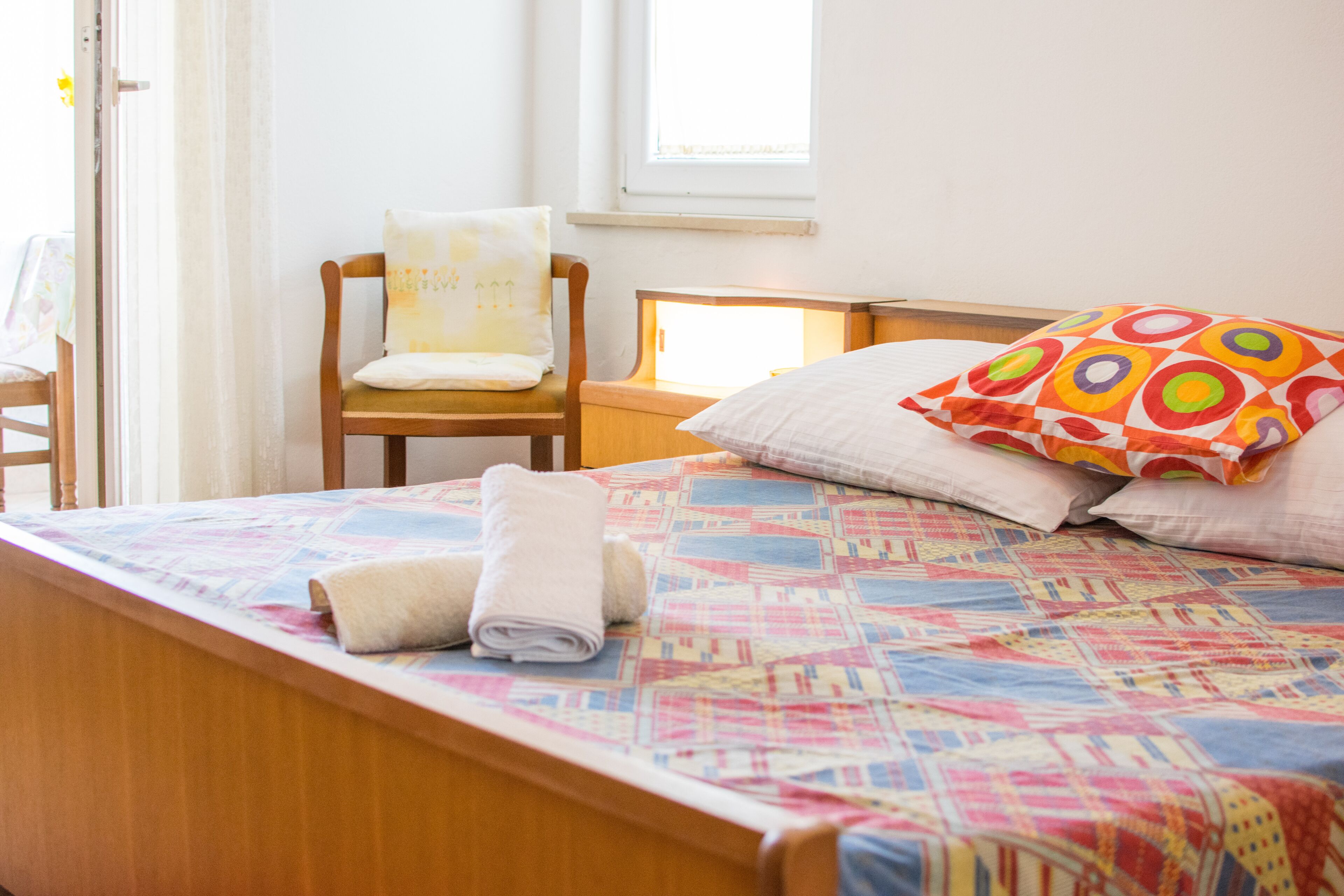 Una cama bien tendida con un colcha de colores, una mesita de noche de madera con una lámpara, una silla con cojín y una ventana con cortinas.