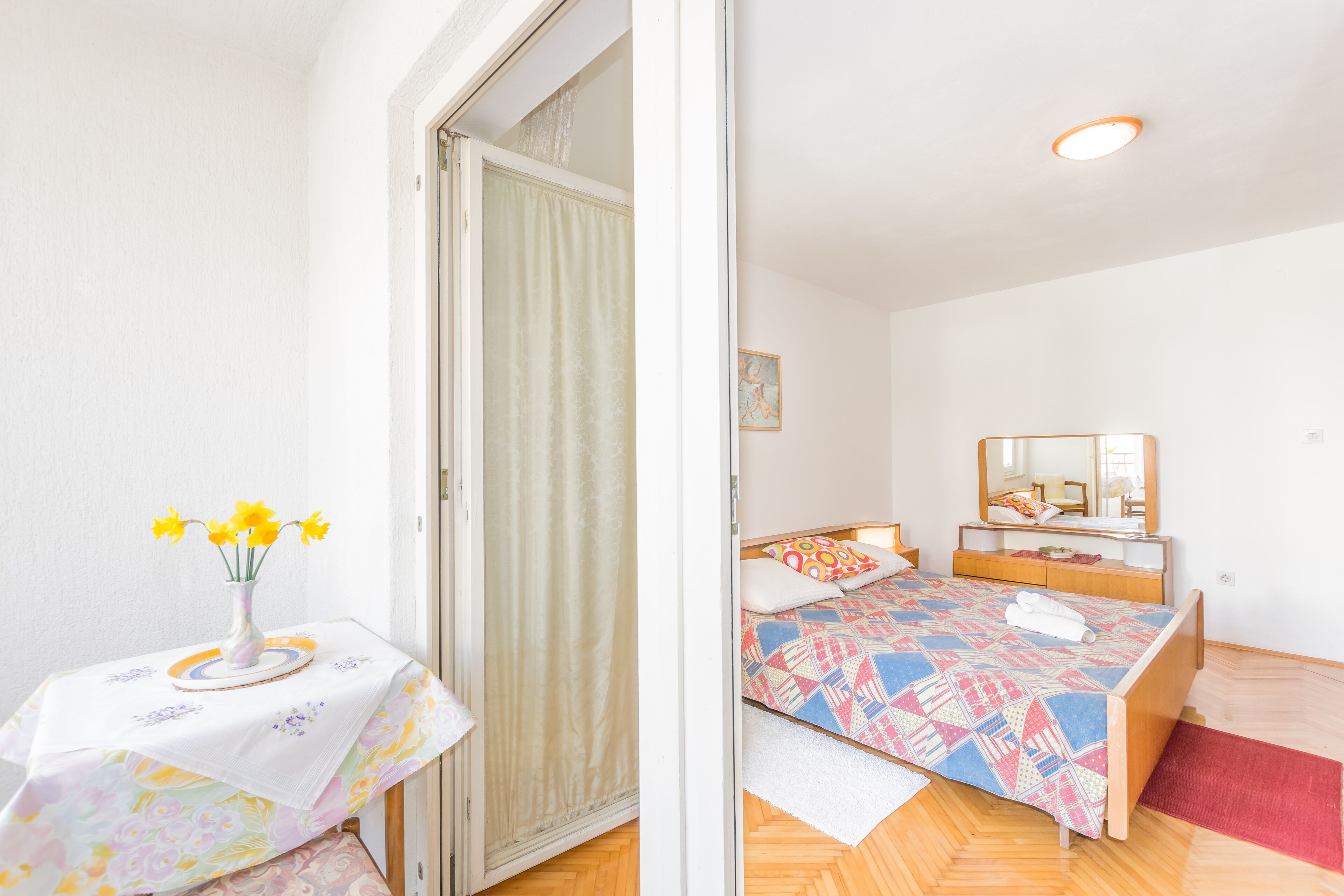 Un dormitorio con cama, una mesita de noche con un florero, un espejo y una puerta que da a otra habitación.