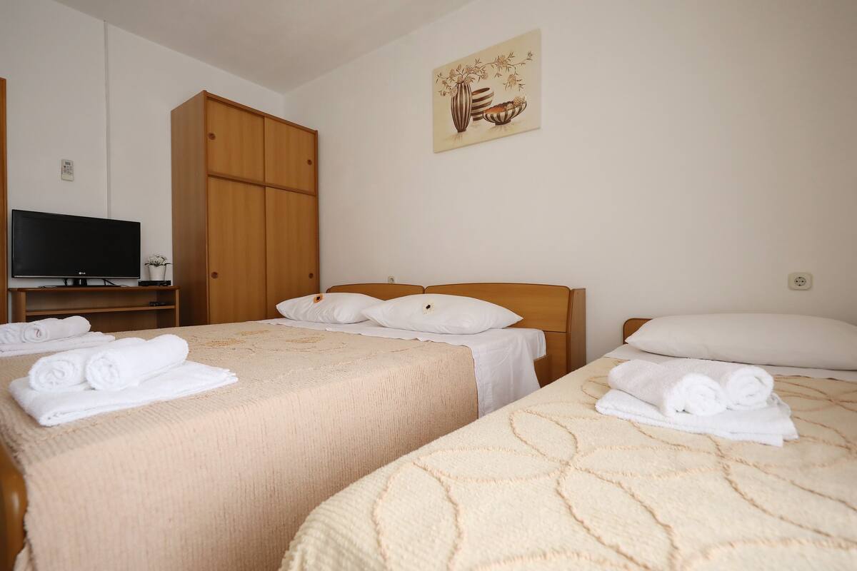 A hotel room with two beds, each with a beige blanket and rolled towels. A television is on a wooden stand to the left.