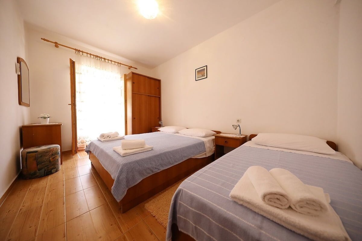 A bedroom with two beds, a wooden floor, a bedside table with a lamp, and a small bedside table with a plant.