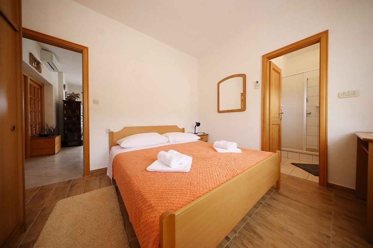 A neatly arranged bedroom with a wooden bed, a bedside table, a mirror, and a bathroom visible through an open door.