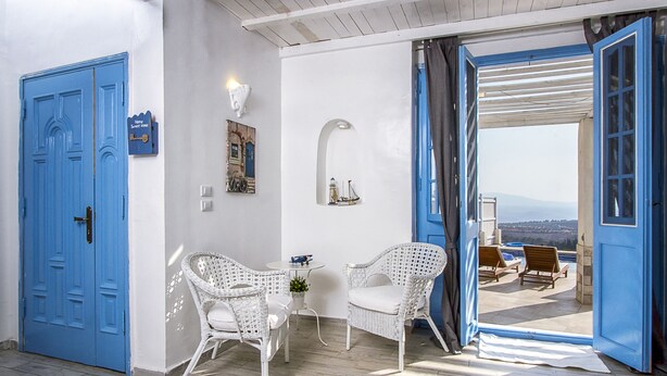 Une pièce avec une porte bleue, des chaises en osier blanches, une petite table et une vue sur l’extérieur.