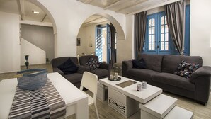 Un salon moderne avec un canapé, un fauteuil, une table basse et une petite table à manger. On aperçoit deux portes bleues et un escalier au fond.