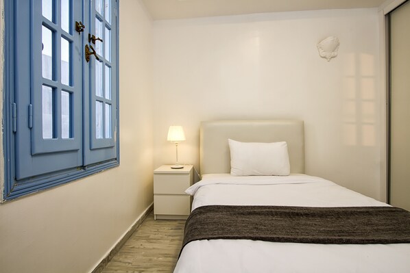 Un dormitorio con cama, mesita de noche, lámpara, ventana con contraventanas azules y un adorno en la pared.