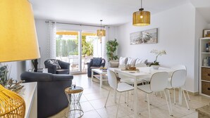 Uma sala de estar moderna com sofá, mesa de centro e área de jantar com cadeiras. Há uma janela grande com cortinas, uma planta e um quadro na parede.