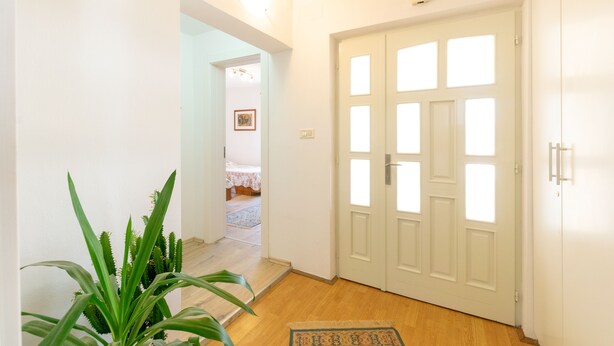 Un couloir au plancher de bois mène à une porte blanche ornée de plusieurs fenêtres. On aperçoit une plante au premier plan.