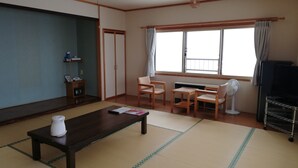 Una sala de estar con esteras de tatami, una mesa de centro de madera oscura, un sofá, un televisor y un ventilador.