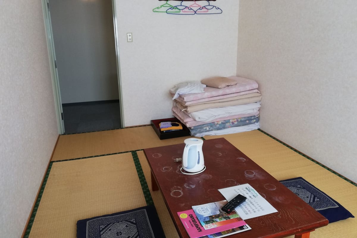 A room with tatami flooring, a wooden table with a kettle and remote control, a stack of folded clothes, and a wall-mounted hanger.