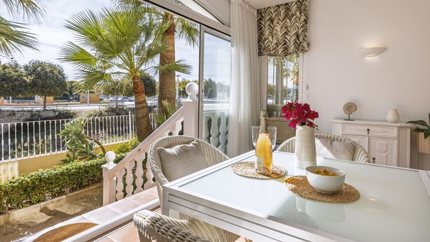 Een eetruimte met een glazen tafel, rieten stoelen, een witte kast en uitzicht op palmbomen en een waterpartij.