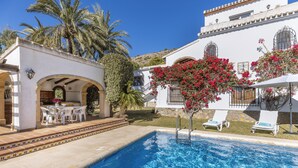 A swimming pool with lounge chairs, a patio area with a table and chairs, and a white building with balconies and red flowers. - Casa Alba (Javea / Xàbia)