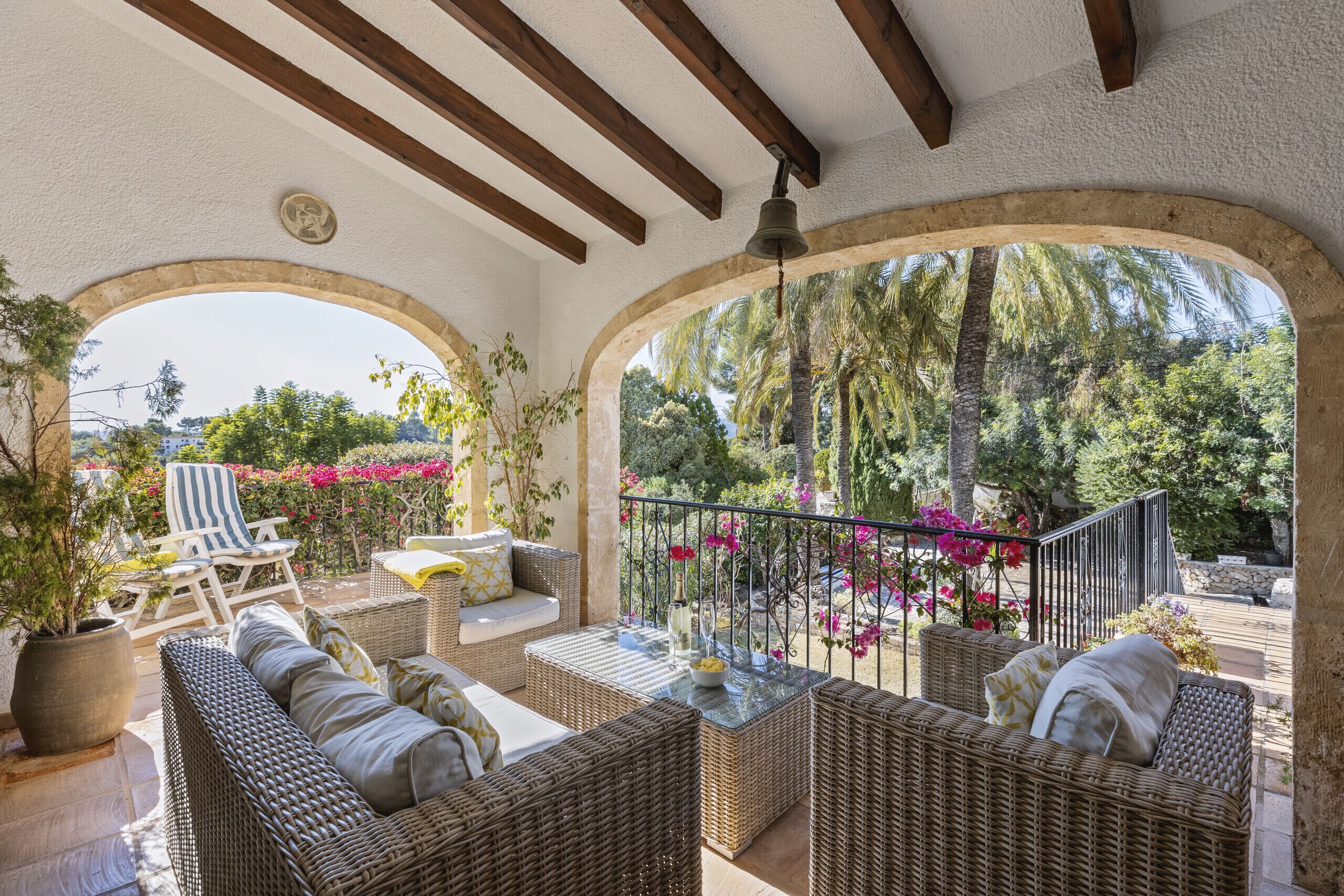 A covered outdoor seating area with wicker furniture, a potted plant, and a view of a garden with palm trees and flowers.