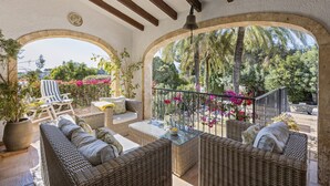 A covered outdoor seating area with wicker furniture, a potted plant, and a view of a garden with palm trees and flowers. - Casa Alba (Javea / Xàbia)